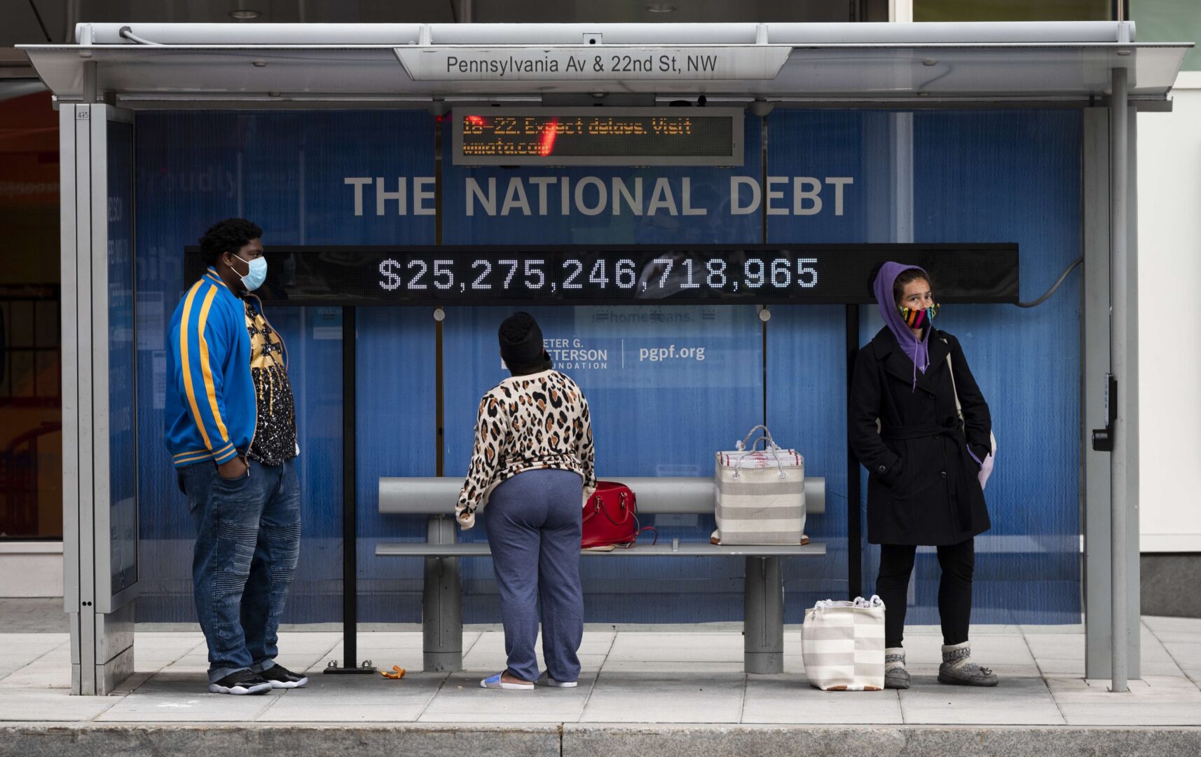 Passengers wearing face masks wait for their bus in front of a national debt display on Pennsylvania Avenue Northwest in Washington on Monday. 